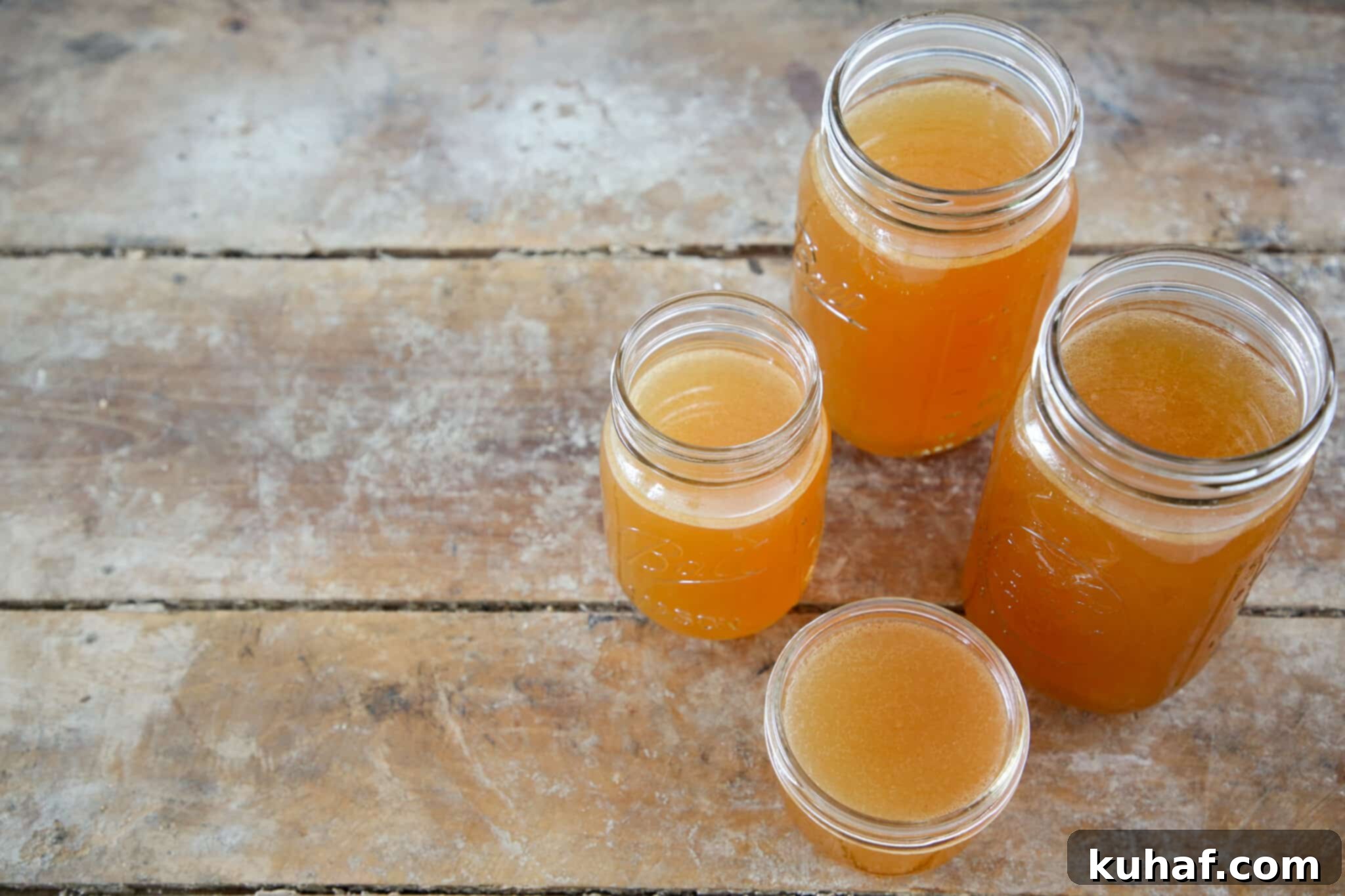 Four jars of homemade chicken stock perfectly lined up on a rustic wood surface, showcasing its golden hue.
