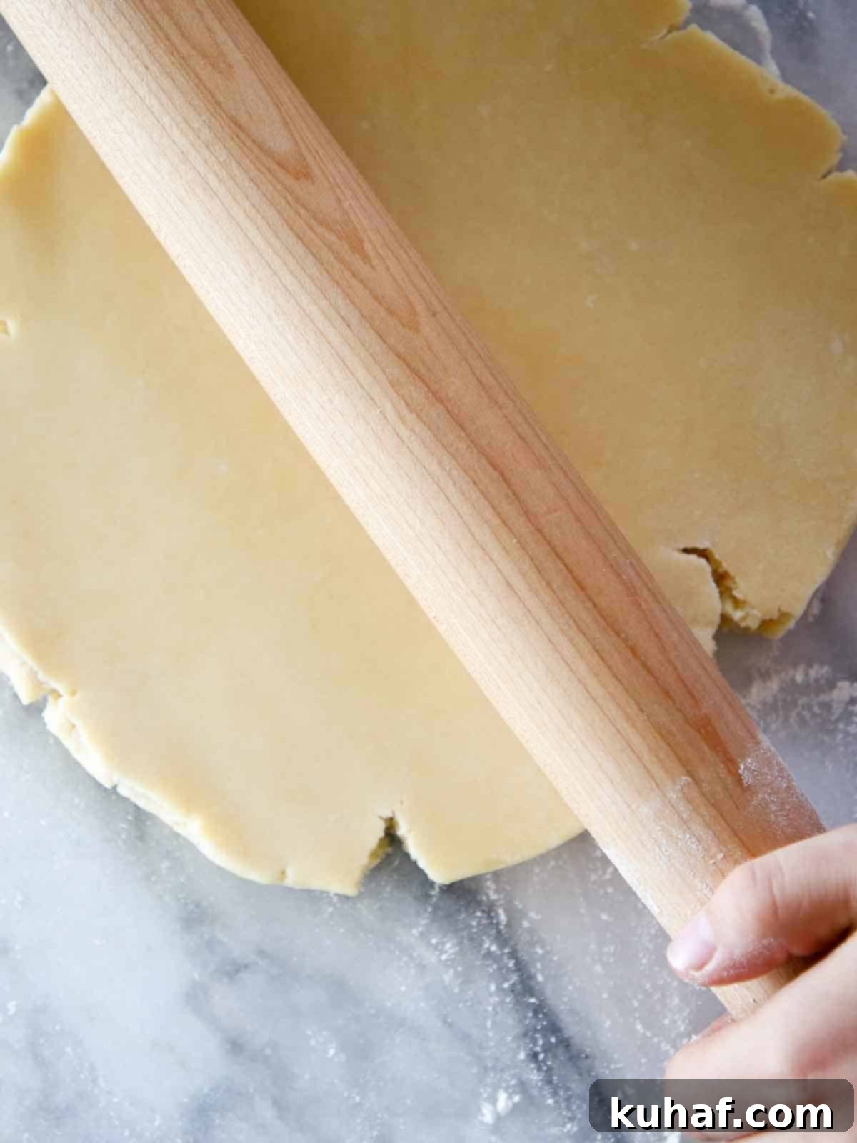 A smooth disk of Pâte Sucrée dough being rolled thin with a wooden rolling pin on a marble surface, preparing it for a tart shell.