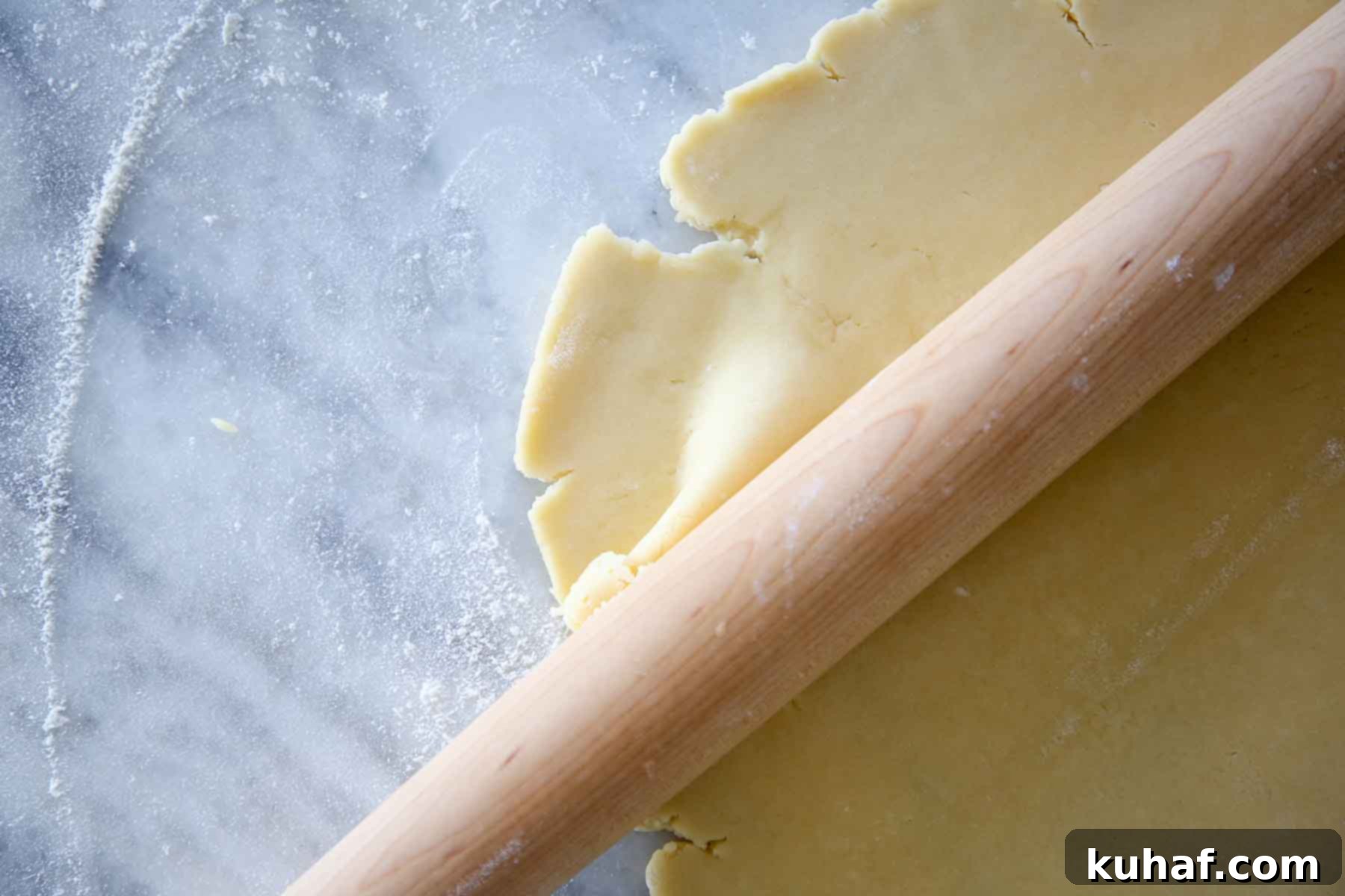 A disk of Pâte Sucrée dough being skillfully rolled thin by a wooden rolling pin on a marble surface, emphasizing its smooth and pliable texture.