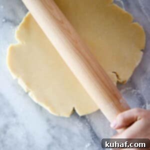 A disk of Pâte Sucrée dough being skillfully rolled by a wooden pin on a marble surface.