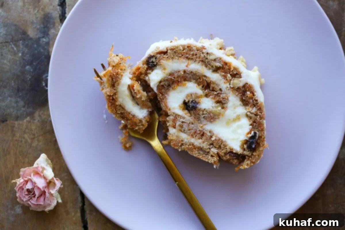 Close-up of a beautifully presented slice of carrot cake roll, revealing the intricate spiral of moist cake, creamy white cream cheese frosting, and visible flecks of carrots, raisins, and walnuts, resting on a white plate.