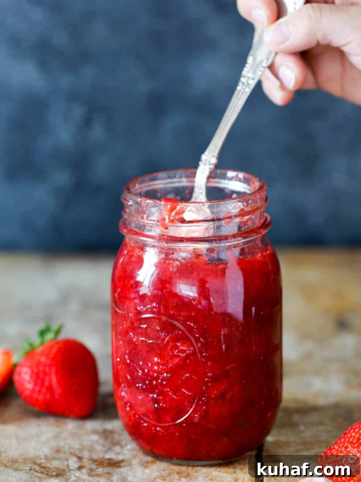 A hand gently dipping a spoon into a jar of freshly made, glistening homemade strawberry jam.