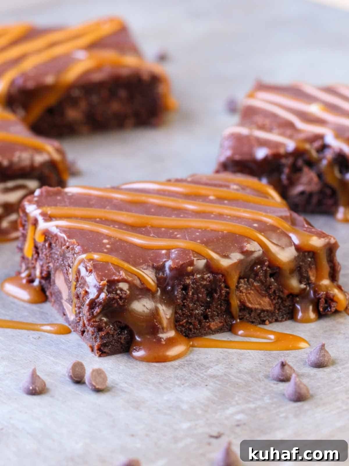 Close-up of a stack of intensely fudgy caramel brownies with visible swirls of salted caramel and a glistening Baileys milk chocolate ganache on top, suggesting a rich, indulgent dessert experience.