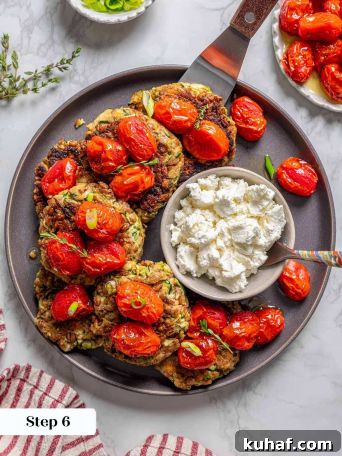 An assembled plate of crispy zucchini fritters, crowned with slow-roasted tomatoes and goat cheese, ready to be served, with a serving spatula beside them.