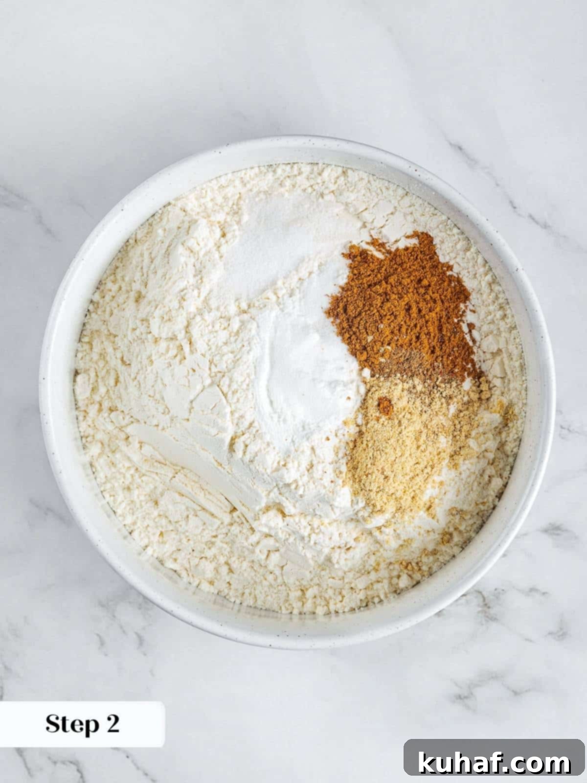 A medium mixing bowl filled with the dry ingredients for pumpkin banana bread: flour, cinnamon, nutmeg, and ginger, ready to be whisked together.