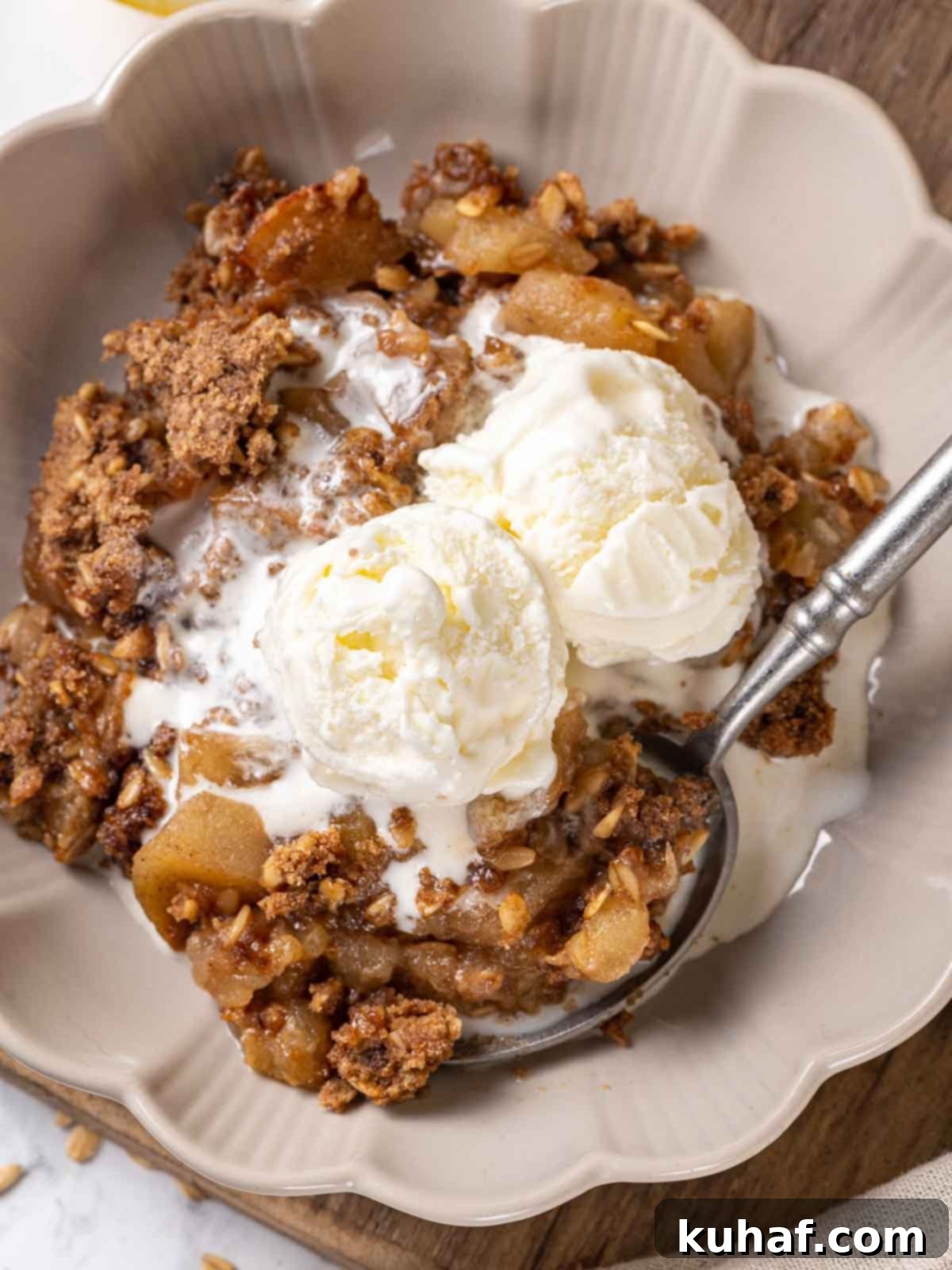 A serving of apple crisp in a bowl, topped with melting vanilla ice cream and a pewter spoon, showcasing its rich texture.