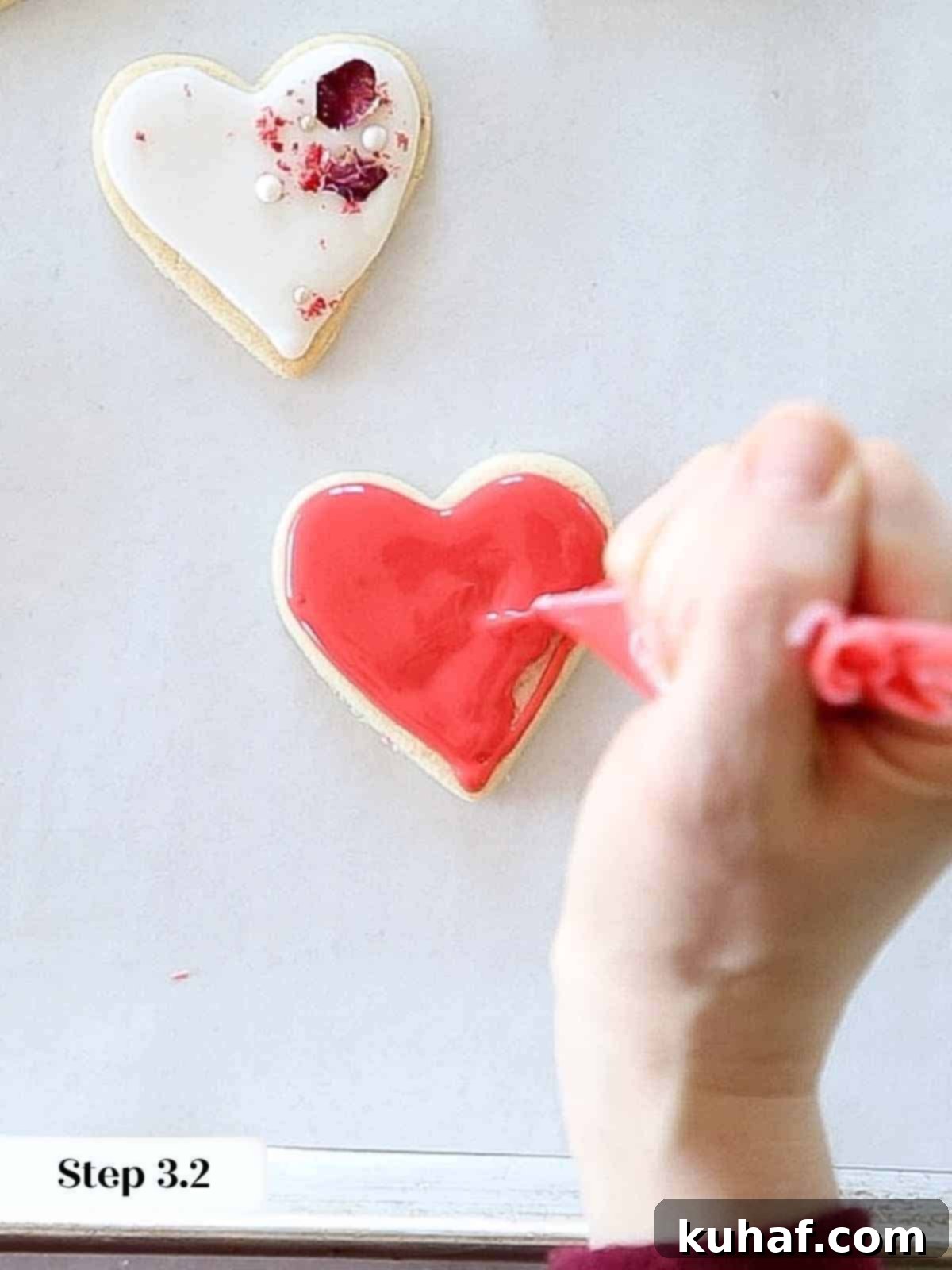 Flooding a heart-shaped sugar cookie with red royal icing, spreading it evenly to the edges for a smooth, vibrant base.