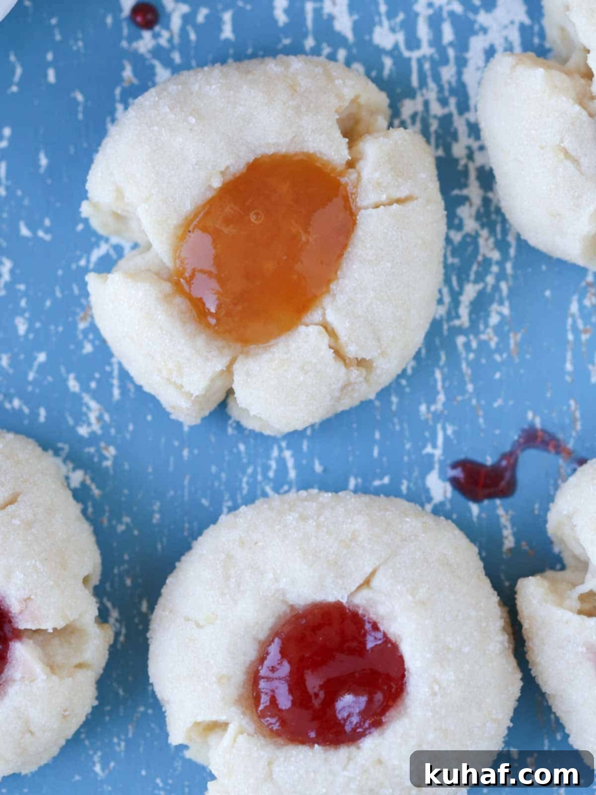Two perfectly baked thumbprint cookies, one with raspberry jam and one with apricot jam, on a blue surface.