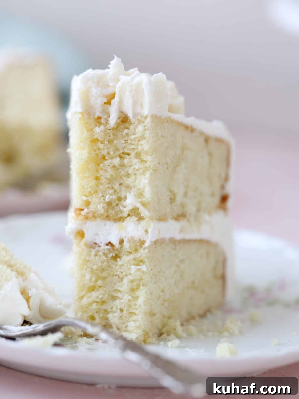 A beautifully sliced moist vanilla cake, showing its delicate, tender crumb and silky frosting layers, standing upright on a plate.