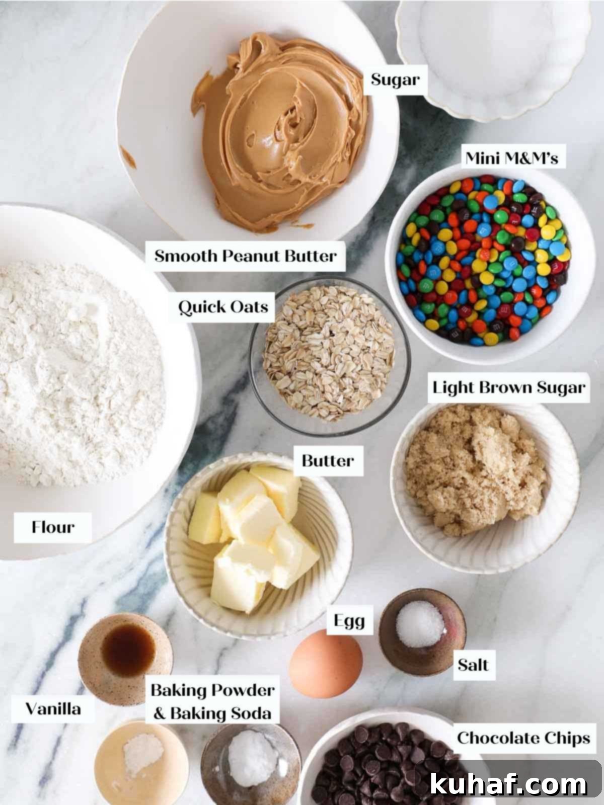 A beautifully arranged flat lay of essential monster cookie ingredients on a pristine white marble counter, featuring oats, creamy peanut butter, and colorful M&M's.