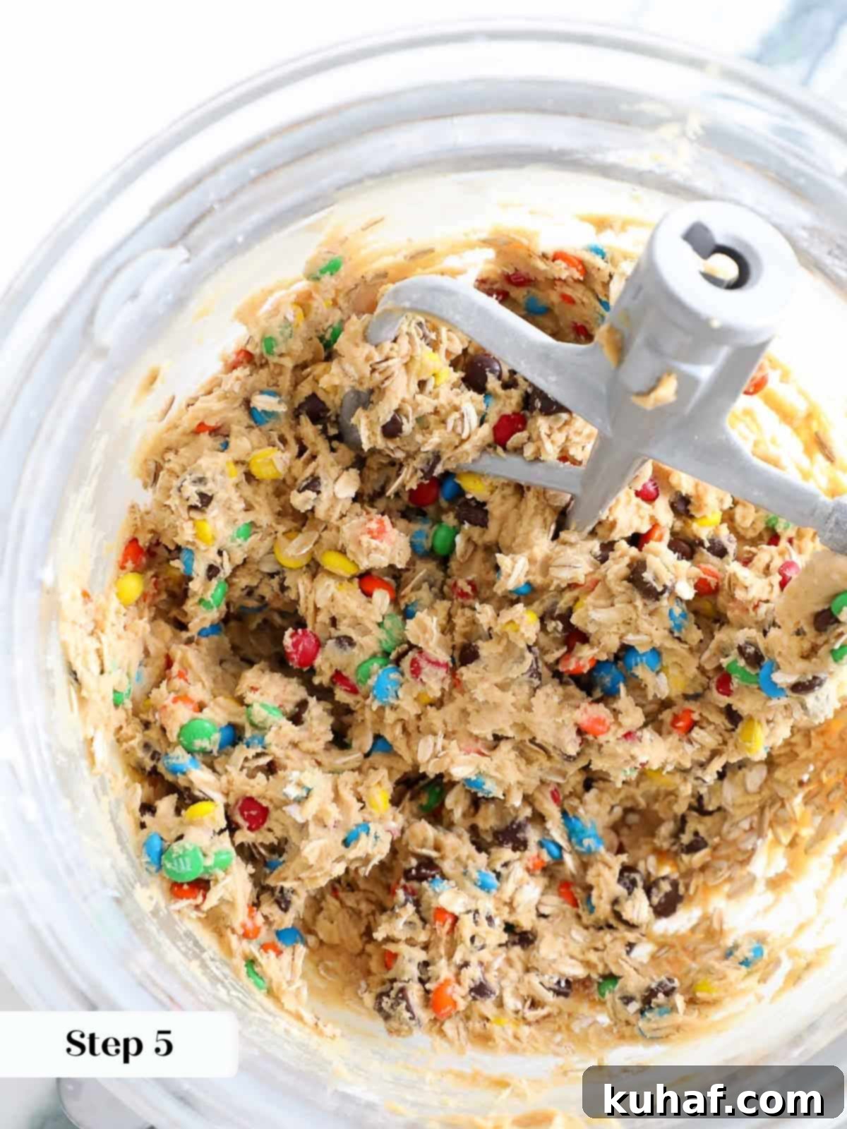 A transparent mixing bowl showcases chunky monster cookie dough, generously packed with a colorful array of mix-ins.
