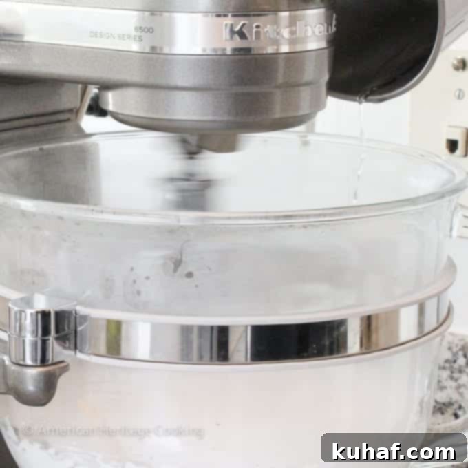 The whisk attachment of a stand mixer, blurred to indicate it's actively mixing at high speed, with a thin stream of sugar syrup pouring down the side of the bowl.
