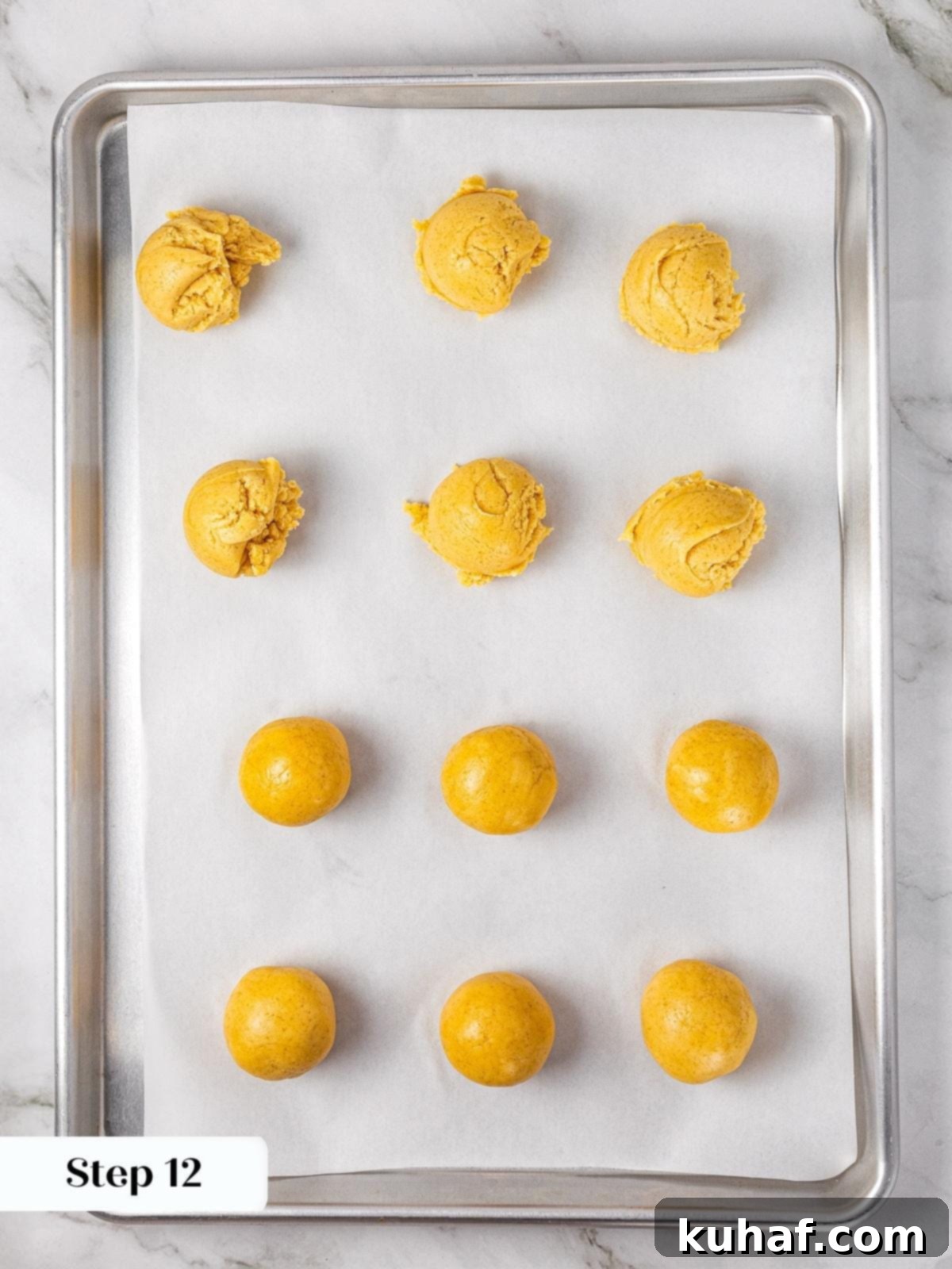 Rows of perfectly scooped apple pie cookie dough balls, each with an indentation, resting on parchment paper on a baking sheet, awaiting their apple filling.