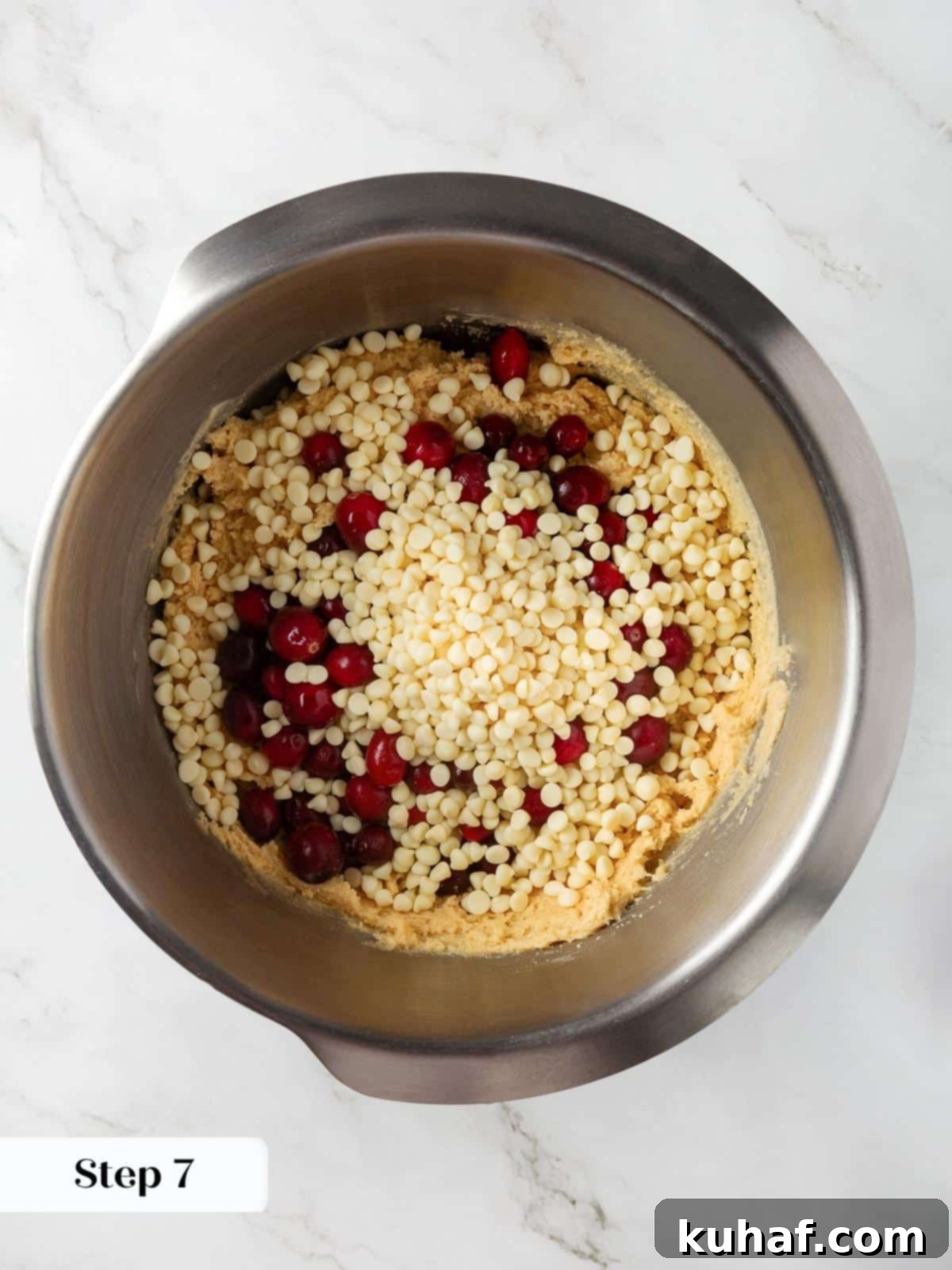 Cranberries and white chocolate chips being folded into thick cookie dough in a mixing bowl.